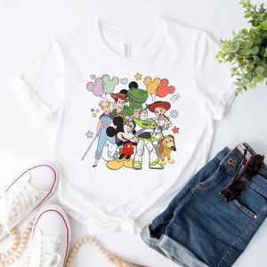 Puede incluir: Camiseta blanca con un gr&aacute;fico colorido de Mickey Mouse, Woody, Buzz Lightyear, Jessie, Rex y algunos globos. El gr&aacute;fico es de estilo de dibujos animados.
