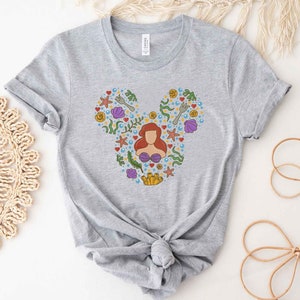 Puede incluir: Camiseta gris con una silueta de Mickey Mouse hecha de coloridas ilustraciones con tem&aacute;tica marina, incluyendo a Ariel de La Sirenita.