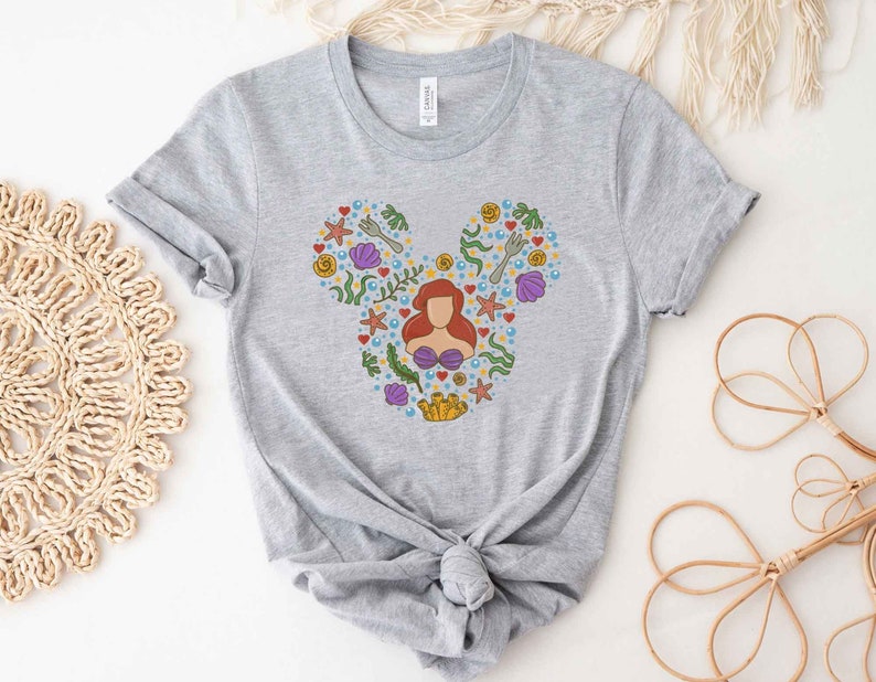 Puede incluir: Camiseta gris con una silueta de Mickey Mouse hecha de coloridas ilustraciones con tem&aacute;tica marina, incluyendo a Ariel de La Sirenita.
