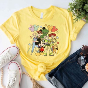 Puede incluir: Camiseta amarilla con un estampado gr&aacute;fico de Mickey Mouse y los personajes de Toy Story Woody, Buzz Lightyear, Jessie y Rex. Los personajes est&aacute;n rodeados de globos y estrellas de colores.