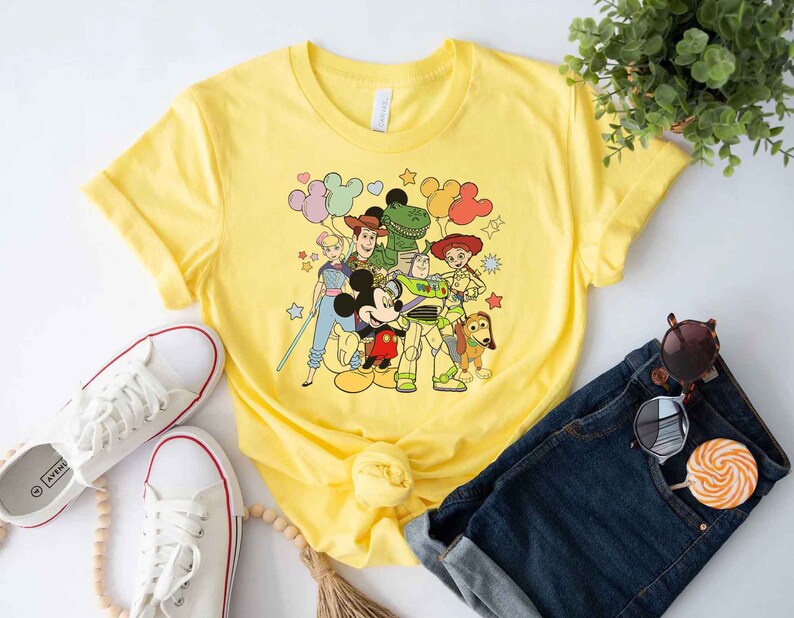 Puede incluir: Camiseta amarilla con un estampado gr&aacute;fico de Mickey Mouse y los personajes de Toy Story Woody, Buzz Lightyear, Jessie y Rex. Los personajes est&aacute;n rodeados de globos y estrellas de colores.