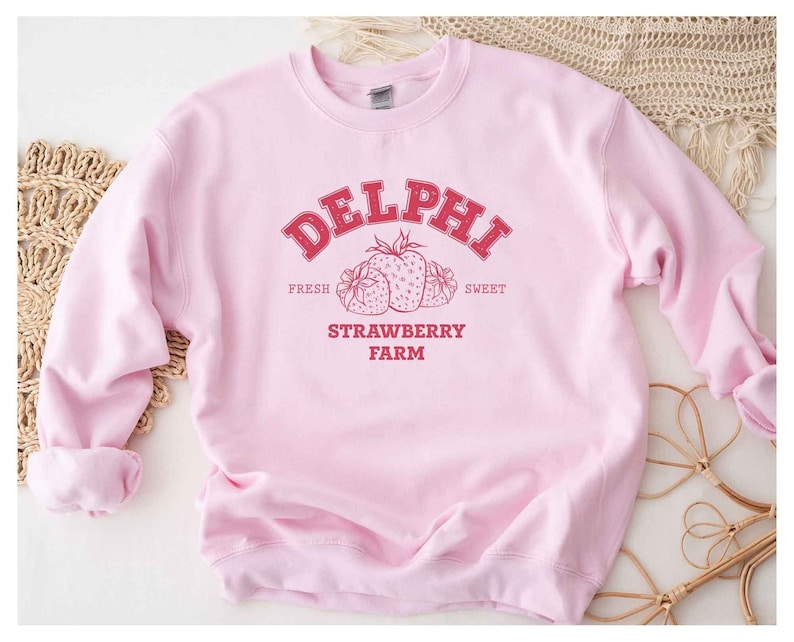 Op de afbeelding: Roze sweatshirt met een rood grafisch ontwerp met de tekst "DELPHI FRESH SWEET STRAWBERRY FARM" en een tekening van drie aardbeien.