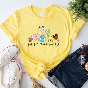May include: Yellow t-shirt with a graphic of a cartoon princess holding a Mickey Mouse ice cream cone and other Disney snacks. The text "BEST DAY EVER" is printed below the graphic.