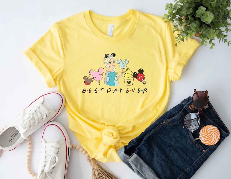 May include: Yellow t-shirt with a graphic of a cartoon princess holding a Mickey Mouse ice cream cone and other Disney snacks. The text "BEST DAY EVER" is printed below the graphic.