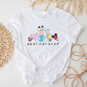 May include: White t-shirt with a graphic of a cartoon character holding various Disney treats. The text "BEST DAY EVER" is printed below the graphic.