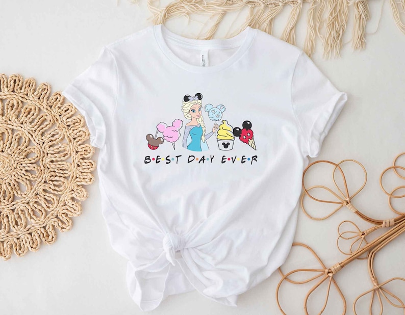 May include: White t-shirt with a graphic of a cartoon character holding various Disney treats. The text "BEST DAY EVER" is printed below the graphic.