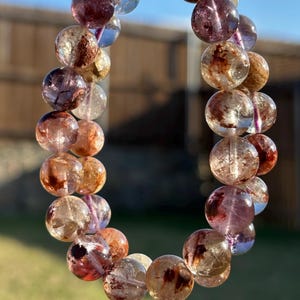 May include: A beaded bracelet composed of translucent, spherical beads in shades of amber, brown, and purple. The beads are strung together, forming a circular shape. The bracelet is displayed against a backdrop of a blue sky and green grass.