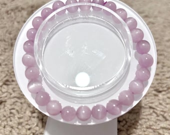 May include: A light purple beaded bracelet displayed on a white and clear acrylic stand. The bracelet features round, polished beads. The stand has a circular top and a rectangular base, set against a neutral-toned background.