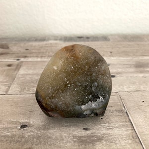 May include: A geode with a rounded shape, featuring a mix of brown, tan, and white colors. The surface is textured with visible crystal formations. The geode is displayed on a wooden surface.
