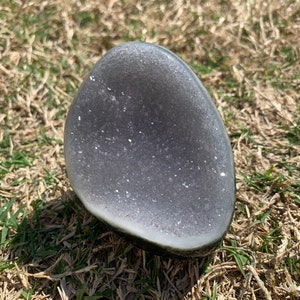 May include: A gray geode stone with a crystalline interior. The stone has a smooth, rounded exterior and a cavity filled with sparkling crystals. The geode is resting on a bed of dry grass and green grass.