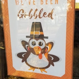 May include: A white sign with orange borders and a brown and white cartoon turkey wearing a black hat with a gold buckle. The sign says "We've Been Gobbled".