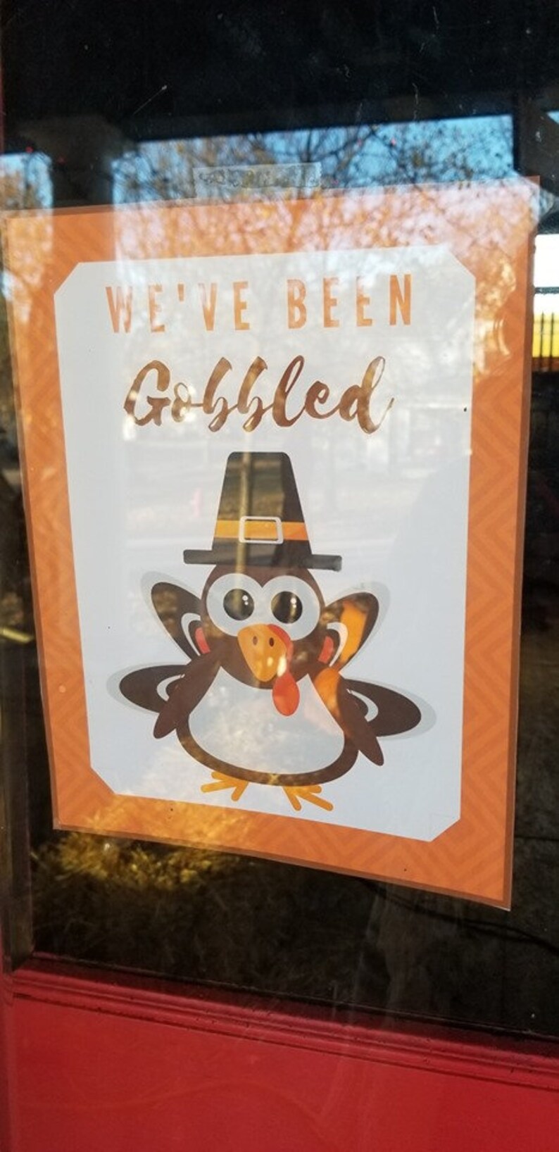 May include: A white sign with orange borders and a brown and white cartoon turkey wearing a black hat with a gold buckle. The sign says "We've Been Gobbled".