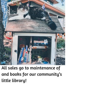 May include: A small wooden library with a cat sitting on the roof. The library has a glass door with a sign that says "Trick or Treat" and is filled with books. The sign below the library says "All sales go to maintenance of and books for our community's little library!"