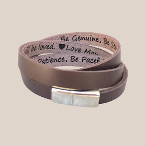 May include: A brown leather wrap bracelet with a silver magnetic clasp. The bracelet is engraved with the words "Be Genuine, Be Strong, will be loved, Love More, Patience, Be Peaceful."
