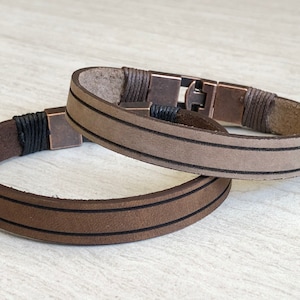 May include: Two leather bracelets with brown and beige leather straps and black stitching. The bracelets have a metal clasp.