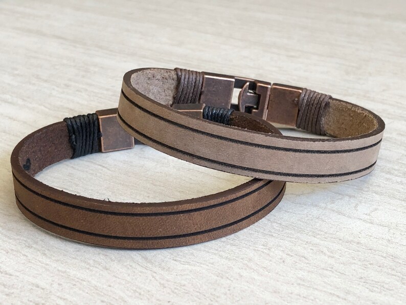 May include: Two leather bracelets with brown and beige leather straps and black stitching. The bracelets have a metal clasp.