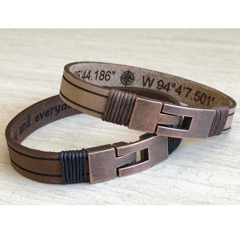 May include: Two leather bracelets with copper clasps. One bracelet has the text "and every" engraved on it. The other bracelet has the text "N 44.186" and "W 94&deg;4'7.501" engraved on it.