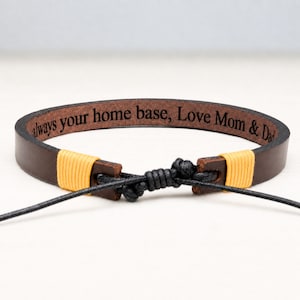 Pode incluir: Uma pulseira de couro castanho escuro com um fecho de cordão preto e nós ajustáveis. A superfície interior da banda de couro está gravada com o texto "always your home base, Love Mom & Dad" e decorada com fio amarelo.