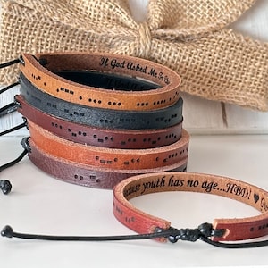 May include: A stack of brown leather bracelets with black text and braille dots. The top bracelet has the text "If God Asked Me To..." and the bottom bracelet has the text "Because youth has no age... HBD!"