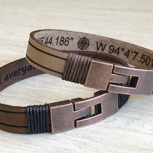 May include: Two leather bracelets with copper clasps. One bracelet is brown with black stitching and the other is beige with brown stitching. The beige bracelet has the coordinates "N 44.186" and "W 94&deg;4'7.501" engraved on it.