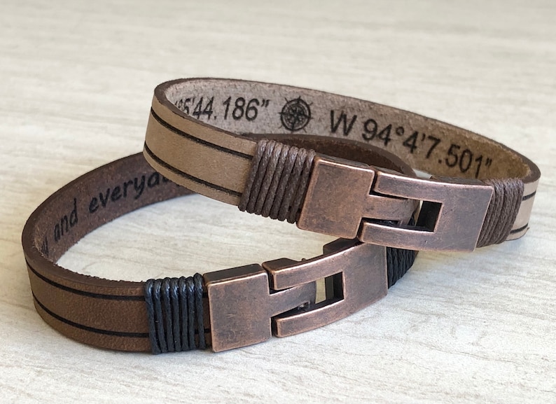 May include: Two leather bracelets with copper clasps. One bracelet is brown with black stitching and the other is beige with brown stitching. The beige bracelet has the coordinates "N 44.186" and "W 94&deg;4'7.501" engraved on it.