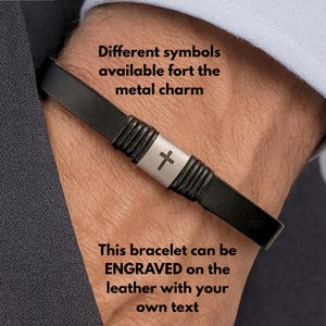 May include: Black leather bracelet with a silver cross charm. The bracelet has a braided design and can be engraved with custom text. The image also includes the text "Different symbols available for the metal charm" and "This bracelet can be engraved on the leather with your own text."