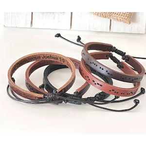 May include: Three leather bracelets with adjustable closures. One bracelet is brown with the text "Be Courageous Joshua 1:9" in black lettering. The other two bracelets are brown and black with braille dots.