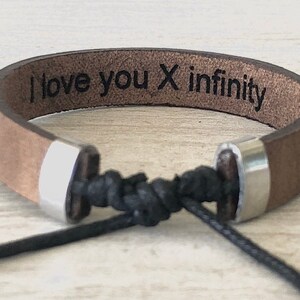 May include: Brown leather bracelet with silver metal accents and a black cord. The bracelet has the text "I love you x infinity" engraved on it.
