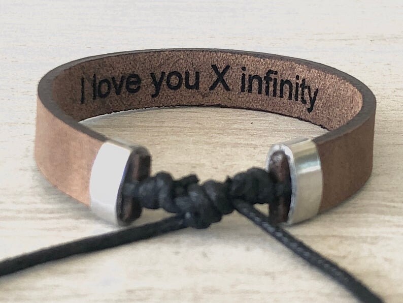 May include: Brown leather bracelet with silver metal accents and a black cord. The bracelet has the text "I love you x infinity" engraved on it.