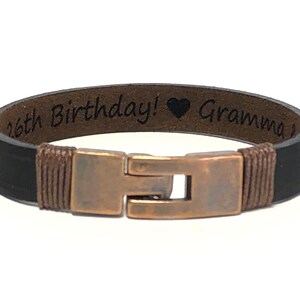 May include: Black leather bracelet with a copper clasp and an engraved message that reads "16th Birthday! &hearts; Grandma".