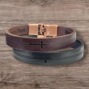 May include: Two leather bracelets with cross designs. One is brown with the text "ma Terri" and the other is black. Both have a copper-colored clasp. The bracelets are displayed on a wooden surface.