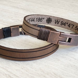 May include: Two brown leather bracelets with copper-toned clasps. The top bracelet has a compass and coordinates engraved: "N 44.186" W 94&deg;4'7.501"