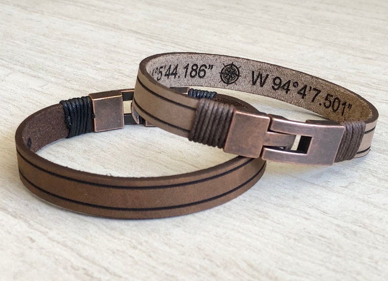May include: Two brown leather bracelets with copper-toned clasps. The top bracelet has a compass and coordinates engraved: "N 44.186" W 94&deg;4'7.501"