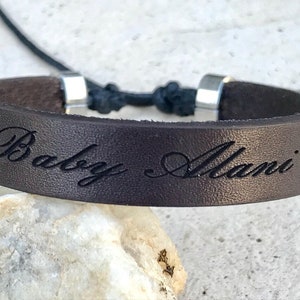 May include: Brown leather bracelet with a silver clasp and the words "Baby Alani" engraved on it, with a heart on each side of the text.