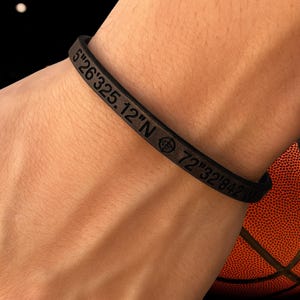 May include: Black bracelet with engraved coordinates: 5°26'325.12"N 72°32'842"W. The bracelet is worn on a wrist, with a basketball visible in the background. The bracelet has a simple, minimalist design.
