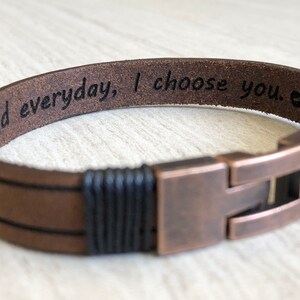 May include: Brown leather bracelet with black braided accents and a copper clasp. The bracelet has the inscription "And everyday, I choose you."