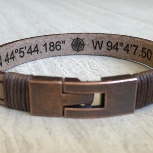 May include: A brown leather bracelet with a copper clasp. The bracelet is engraved with the coordinates "N 44&deg; 5' 44.186" and "W 94&deg; 4' 7.501" and a compass symbol.