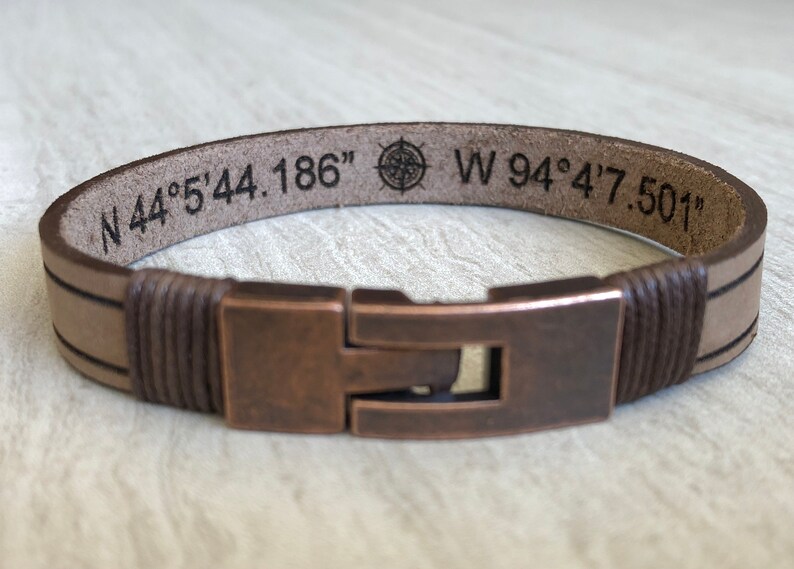 May include: A brown leather bracelet with a copper clasp. The bracelet is engraved with the coordinates "N 44&deg; 5' 44.186" and "W 94&deg; 4' 7.501" and a compass symbol.