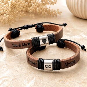 May include: Two brown leather bracelets with adjustable black cord closures. One bracelet features a silver-toned metal bead with a black heart and the text You & Me. The second bracelet has a silver-toned bead with an infinity symbol and the text Forever Always.