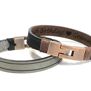 May include: Two leather bracelets with copper clasps. One bracelet is black with a brown leather band and the text "Happy Birthday! &hearts; Grandma" engraved on it. The other bracelet is gray with two white stripes.