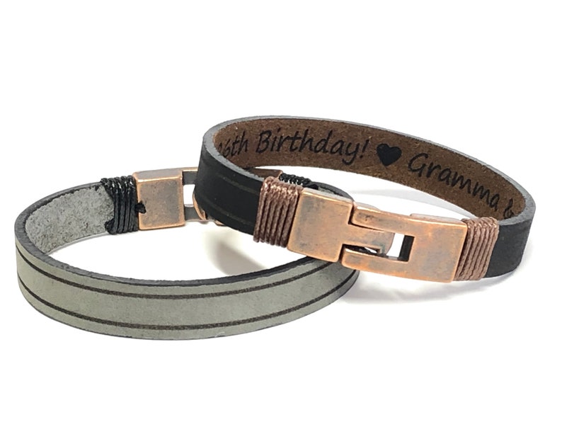 May include: Two leather bracelets with copper clasps. One bracelet is black with a brown leather band and the text "Happy Birthday! &hearts; Grandma" engraved on it. The other bracelet is gray with two white stripes.
