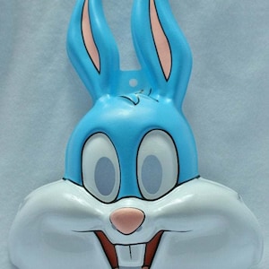 Könnte beinhalten: Eine hellblaue Bugs Bunny-Maske mit großen weißen Augen, einer rosa Nase und einem breiten, zahnigen Grinsen. Die Maske hat lange, aufrechte Ohren mit rosa Innendetails. Ein lustiger Artikel zum Thema Cartoon.