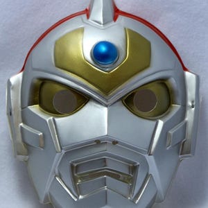 Può includere: Una maschera argentata e dorata con una gemma blu e dettagli rossi. La maschera presenta un design futuristico con fori per gli occhi e una parte superiore a punta. Per cosplay o costumi.