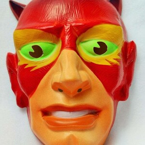 May include: A devil mask with a red head and horns, yellow and red accents around the eyes, and a light orange face. The eyes are green with brown pupils, and the mouth is open in a slight grin.