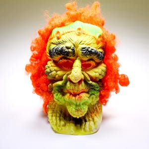 Puede incluir: Una máscara grotesca con una cara amarilla y verde, cuencas de ojos negras y una nariz larga y puntiaguda. La máscara está rematada con cabello rizado de color naranja brillante. El diseño general sugiere un monstruo o criatura, posiblemente para Halloween o disfraces.
