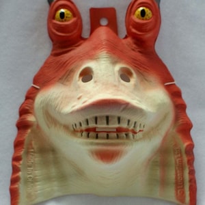 Könnte beinhalten: Eine Kunststoffmaske mit der Figur Jar Jar Binks aus Star Wars. Die Maske ist hauptsächlich orange und cremefarben, mit großen gelben Augen und einem breiten Grinsen. Sie hat ein Loch zum Aufhängen und den Text "Made in China" und "Lucasfilm Ltd."