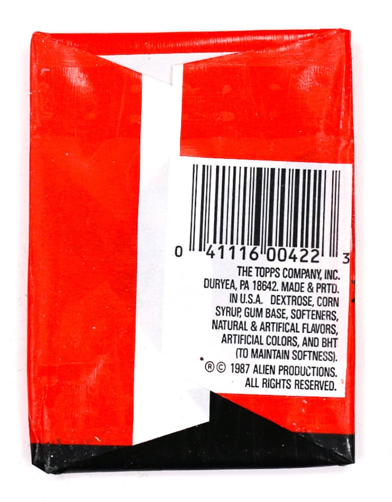 May include: A red and white package with a barcode and text that reads "THE TOPPS COMPANY, INC. DURYEA, PA 18642. MADE & PRTD. IN U.S.A. DEXTROSE, CORN SYRUP, GUM BASE, SOFTENERS, NATURAL & ARTIFICAL FLAVORS, ARTIFICIAL COLORS, AND BHT (TO MAINTAIN SOFTNESS). &copy; 1987 ALIEN PRODUCTIONS. ALL RIGHTS RESERVED."