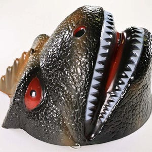 Könnte beinhalten: Eine Maske eines Dinosauriers aus geformtem Kunststoff mit einer strukturierten, dunkelgrünen und braunen Außenseite. Die Maske hat rote Augen, einen weit geöffneten Mund mit weißen Zähnen und eine braune Flosse an der Seite. Ein lustiges Kostümzubehör.