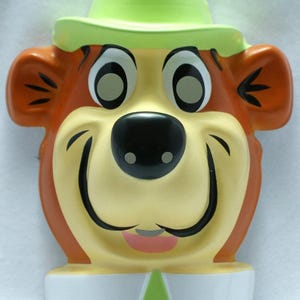 Pode incluir: Uma decoração de parede em cerâmica com um personagem de urso de desenho animado. O urso tem um rosto amarelo claro, orelhas castanhas e um nariz preto. Ele usa um chapéu verde e uma gola branca com um triângulo verde. Os olhos do urso são grandes e pretos com reflexos brancos.