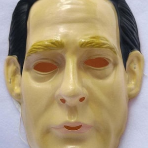 May include: A full-face mask with a realistic human face. The mask is primarily a pale yellow color, with black hair, yellow eyebrows, and orange eye holes. The mouth is slightly open, and the mask has a smooth finish.